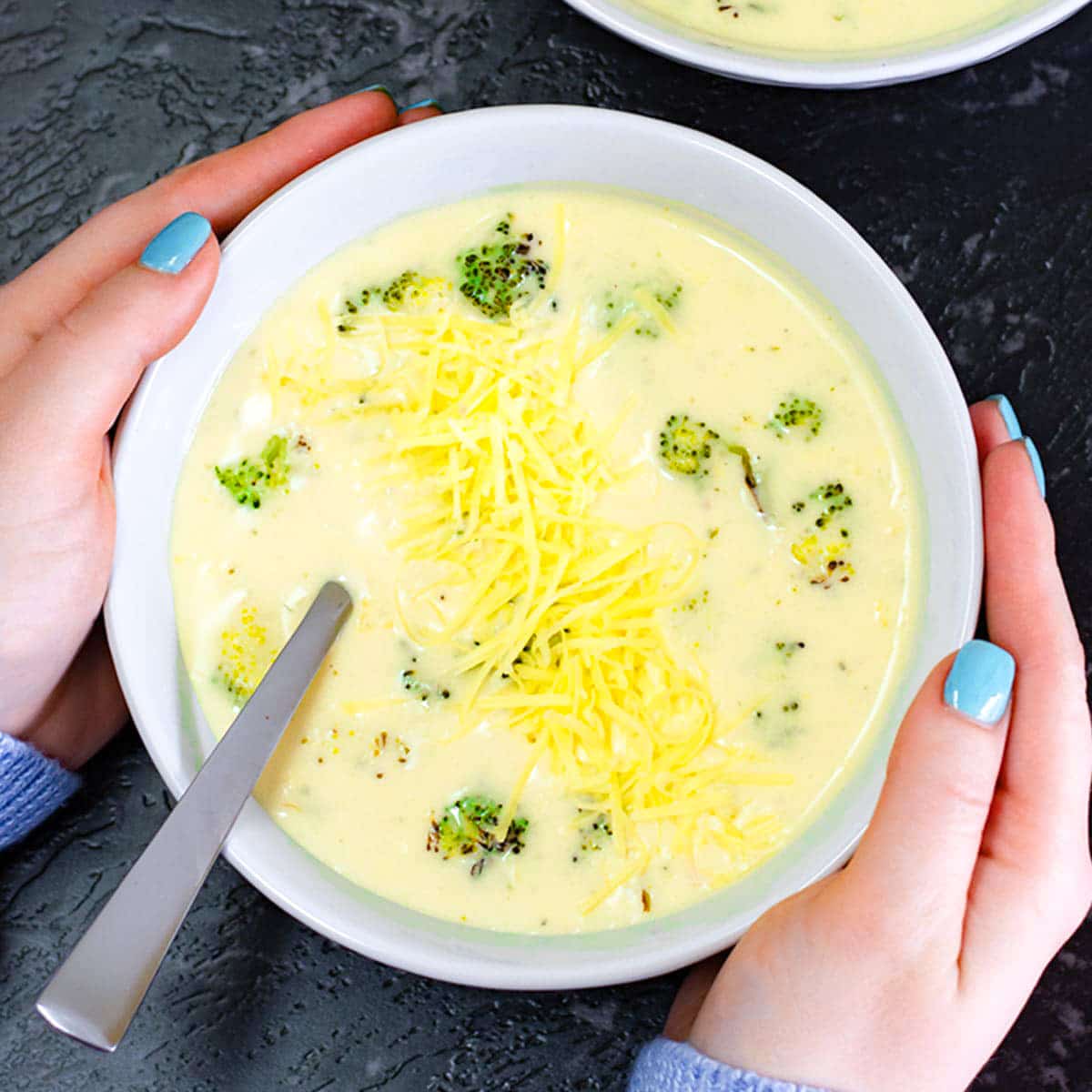 Keto broccoli cheese soup in a white bowl with a silver spoon being held by two hands.