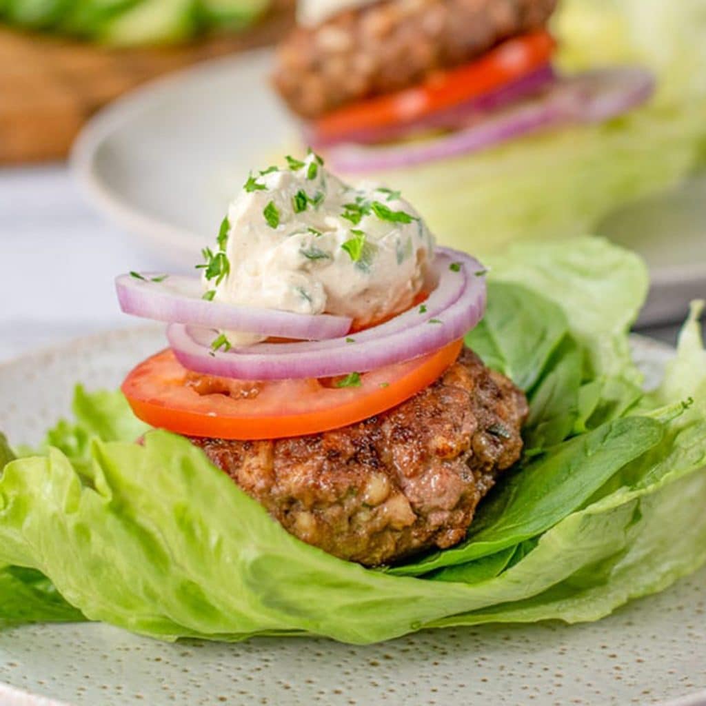 Keto lamb burger patty on a lettuces leaf topped wit a slice of tomato and cucumber sauce.
